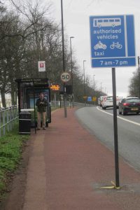 Picture of the shared pavement southbound from Gosforth alongside the Great North Road showing a man waiting at a bus stop and signs and a lamppost in the middle of the pavement.