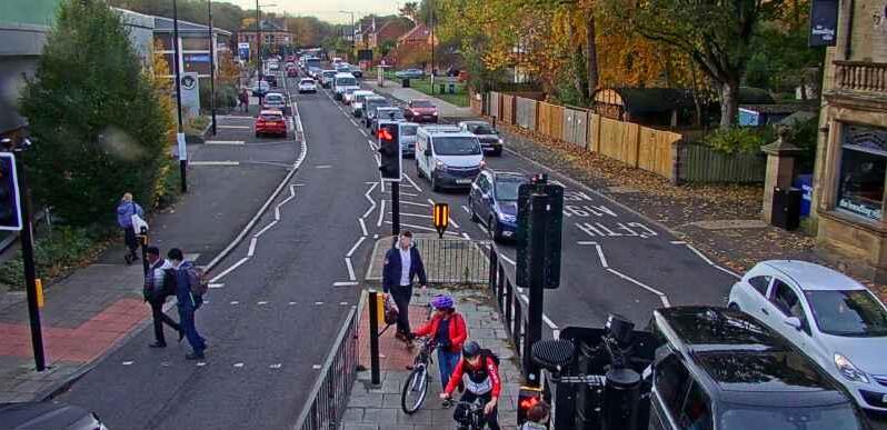 The picture shows a queue of traffic and a pedestrian crossing with people walking and two people with bikes.