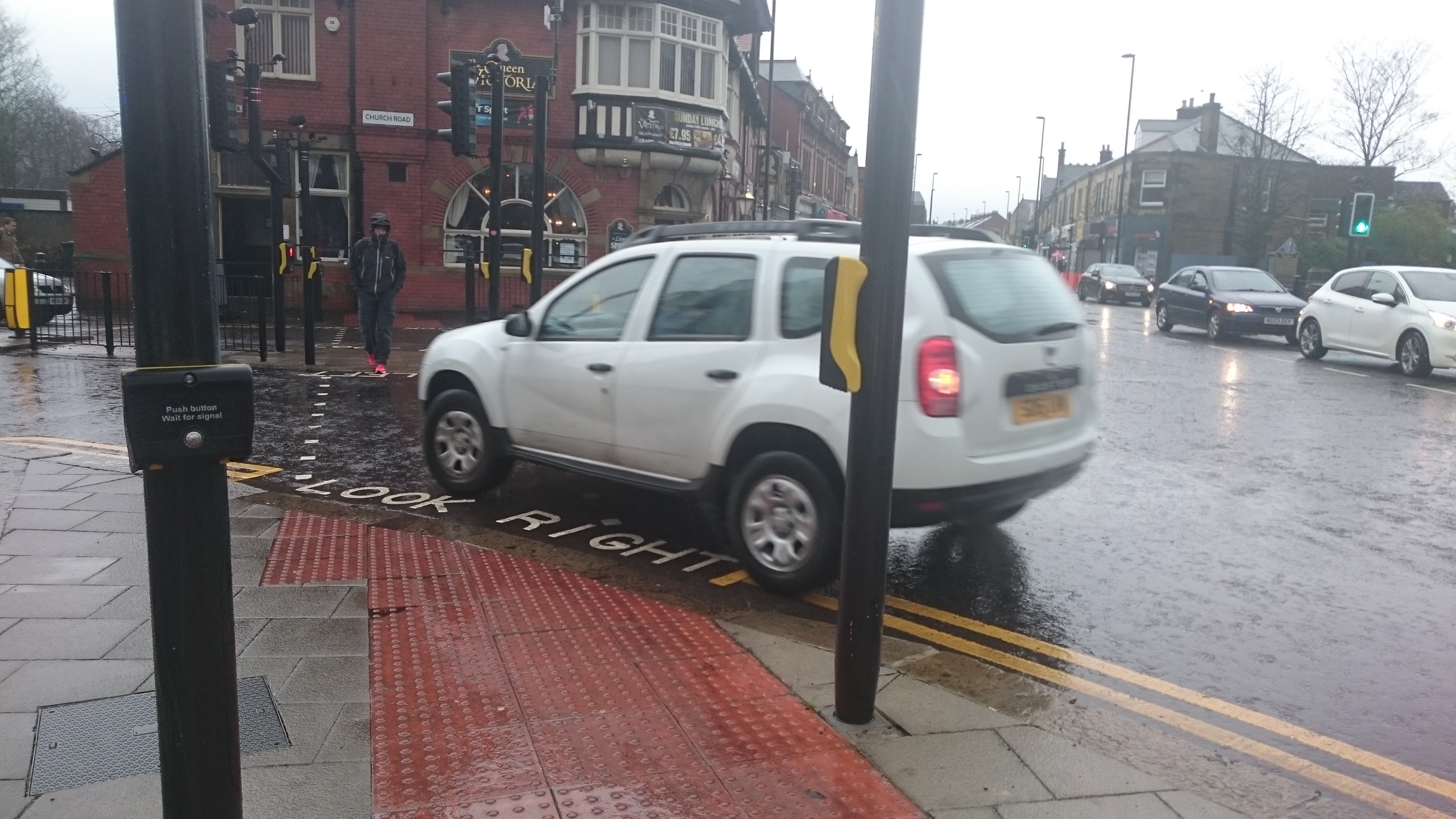 image of crossing across church road heading towards the Queen Victoria pub. Showing the tactile paving and the curved edge of the pavment. Look right is painted on the road. A car is turning across the crossing and is inches away from the pavement edge