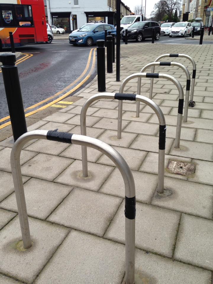 Five silver coloured bicycle racks outside of the gosforth hotel. Each bike rack has about eight inches of black tape wrapped around the middle of the top and front edges.