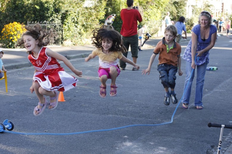 Three children jumping through a skipping rope in the street. Adults in the background.