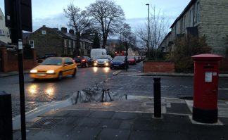 Photo of junction of Ivy Road with Salters Road. Vehicles are travelling up Salters with queued vehicles all the way back from the high street. In the foreground is a post box and pillar, with a deep puddle at the kerbside.