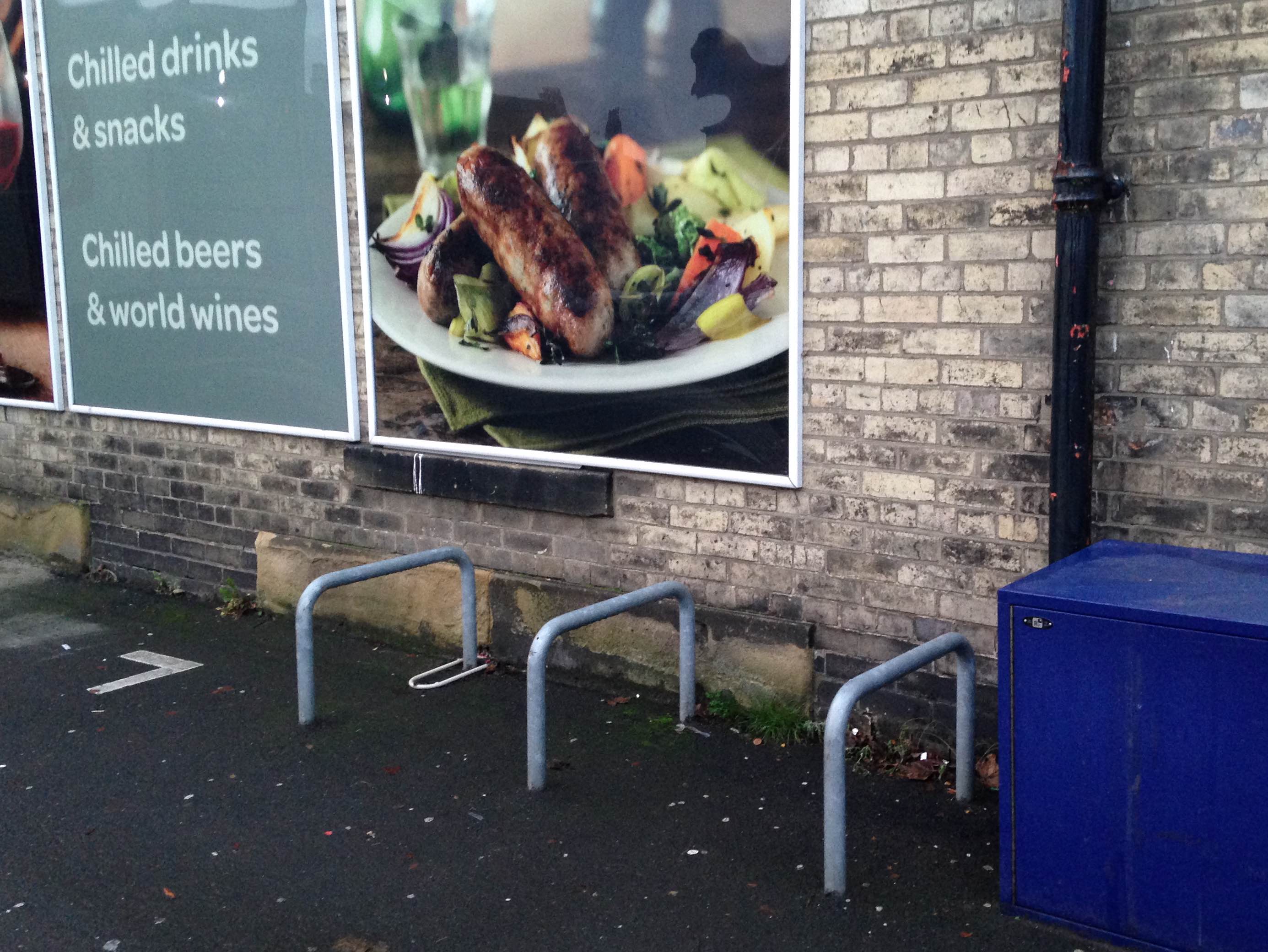 Photograph of three metal bike racks about 3 foot high lined up close to the shop wall with a blue utilities box to the right.