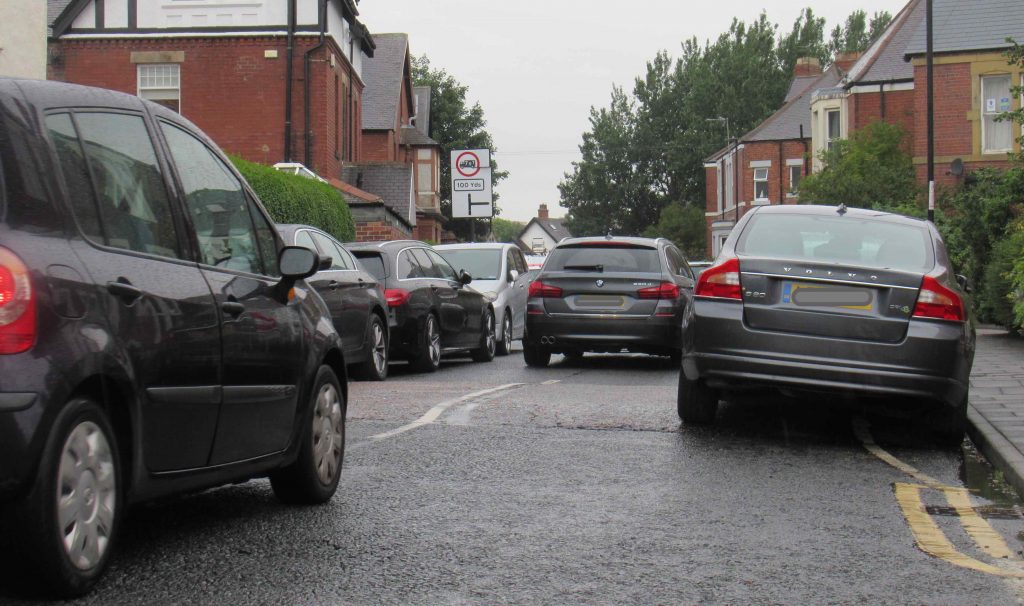 Pircture of cars on Stoneyhurst Road in single file in between two rows of parked cars.