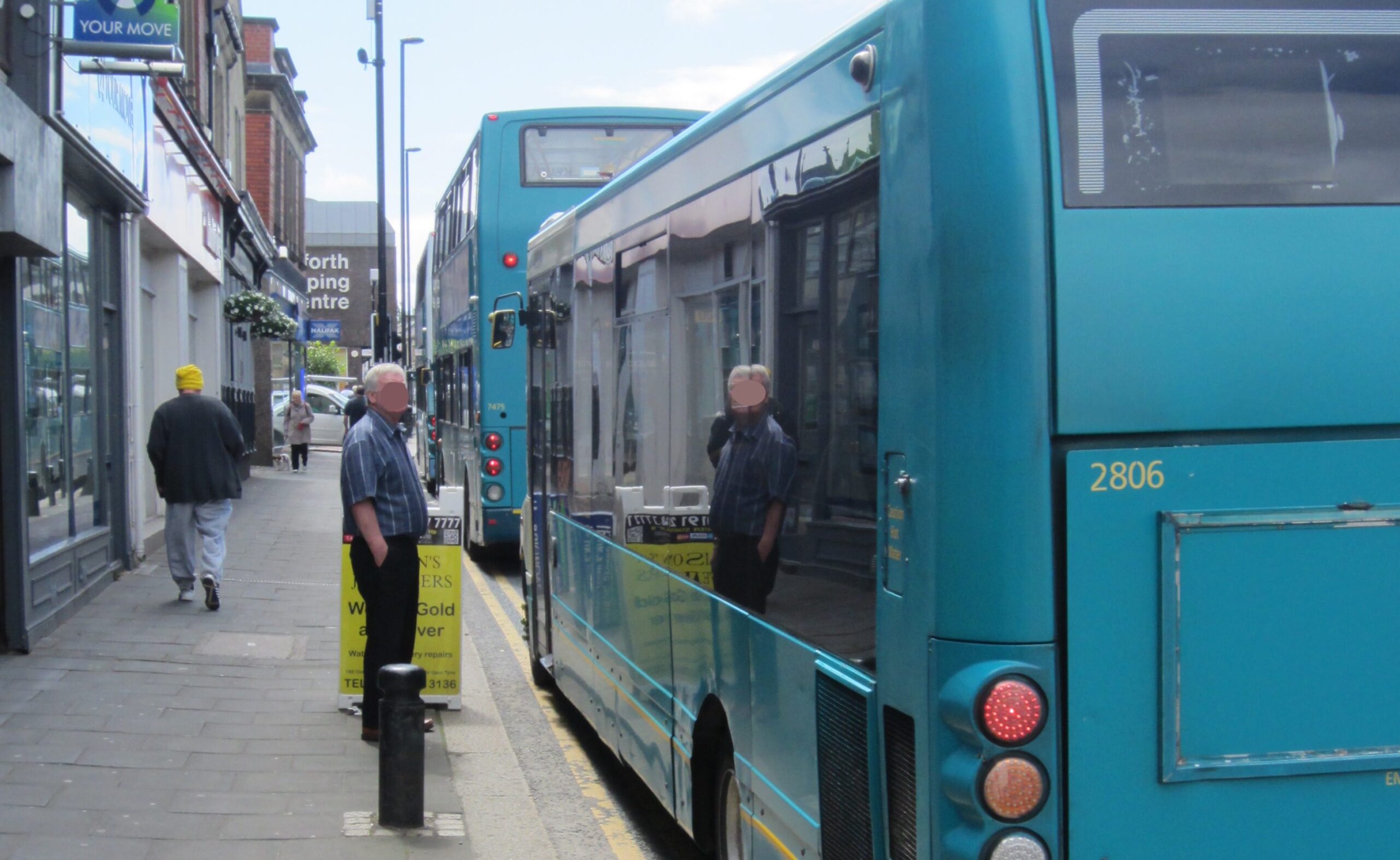 Man standing on the edge of the pavement on Gosforth High Street next to a bus.